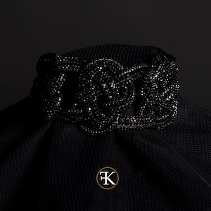 Peut inclure: Un collier ras-du-cou noir doté d'un motif central en forme de nœud incrusté de nombreuses petites perles noires scintillantes. La pièce est présentée sur un fond en tissu noir et comporte un logo circulaire avec les lettres FK en blanc.