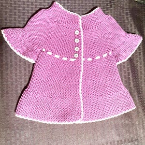 May include: A pink knitted baby cardigan with white trim and buttons. The cardigan has a scalloped edge and a slightly flared shape.