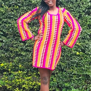 Puede incluir: Un vestido de crochet con rayas rosas, naranjas y blancas. El vestido tiene mangas largas y un corte holgado.