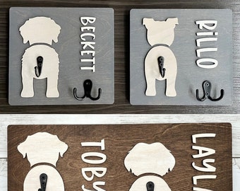 May include: A wooden dog leash holder with four hooks. The holder has a brown wood background with white dog silhouettes and names: Beckett, Pillo, Toby, and Layla.