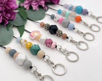 May include: A set of colorful beaded lanyards with keychains. Each lanyard features a different pattern of beads, including rainbow, floral, and geometric designs. The lanyards are made with a variety of materials, including wood, silicone, and acrylic.