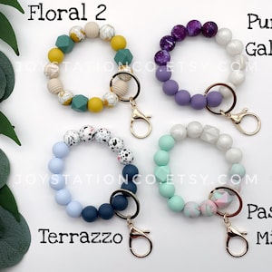 May include: Four colorful beaded keychains with gold key rings. The keychains are made of silicone beads in various patterns and colors, including floral, terrazzo, purple galaxy, and pastel mint.