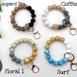 May include: Four colorful beaded keychains with gold key rings. The keychains are made of different colored beads, including tan, black, white, gray, blue, and yellow. The keychains are labeled "Leopard Tan", "Coffee", "Floral 1", and "Surf".