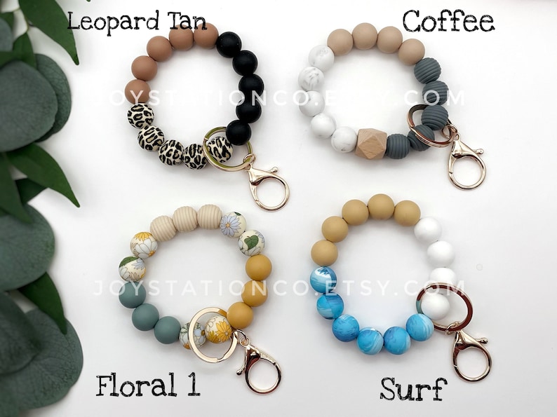 May include: Four colorful beaded keychains with gold key rings. The keychains are made of different colored beads, including tan, black, white, gray, blue, and yellow. The keychains are labeled "Leopard Tan", "Coffee", "Floral 1", and "Surf".