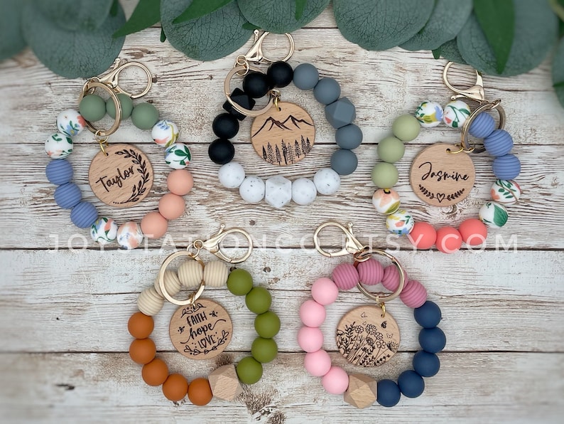May include: Five colorful silicone beaded keychains with wooden charms. The charms feature personalized names, a mountain scene, a floral design, and the words "Faith, Hope, Love".