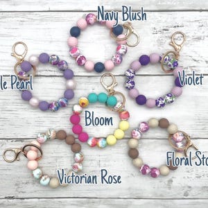 May include: Ten colorful silicone beaded keychains with floral patterns and gold key rings. The keychains are labeled with names like "Navy Blush", "Purple Pearl", "Violet", "Bloom", "Victorian Rose", and "Floral Stone".