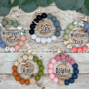 May include: Assortment of colorful silicone beaded keychains with wooden charms. Each keychain features a different color combination and a personalized wooden charm with a name or word. The charms include "Wildflower Peach", "Wildflower Coral", "Slate", "Vino", and "Dolphin".