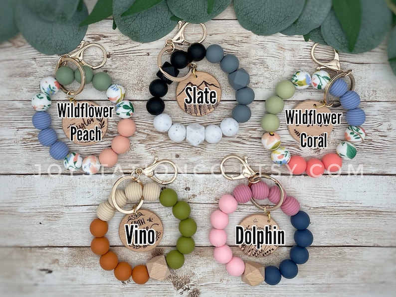 May include: Assortment of colorful silicone beaded keychains with wooden charms. Each keychain features a different color combination and a personalized wooden charm with a name or word. The charms include "Wildflower Peach", "Wildflower Coral", "Slate", "Vino", and "Dolphin".