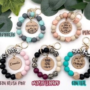 May include: Assortment of colorful beaded keychains with wooden charms. The charms feature various phrases, including "Boy Mom", "Faith Hope Love", "Teach Love Inspire", "Be the Change", and "Danielle".