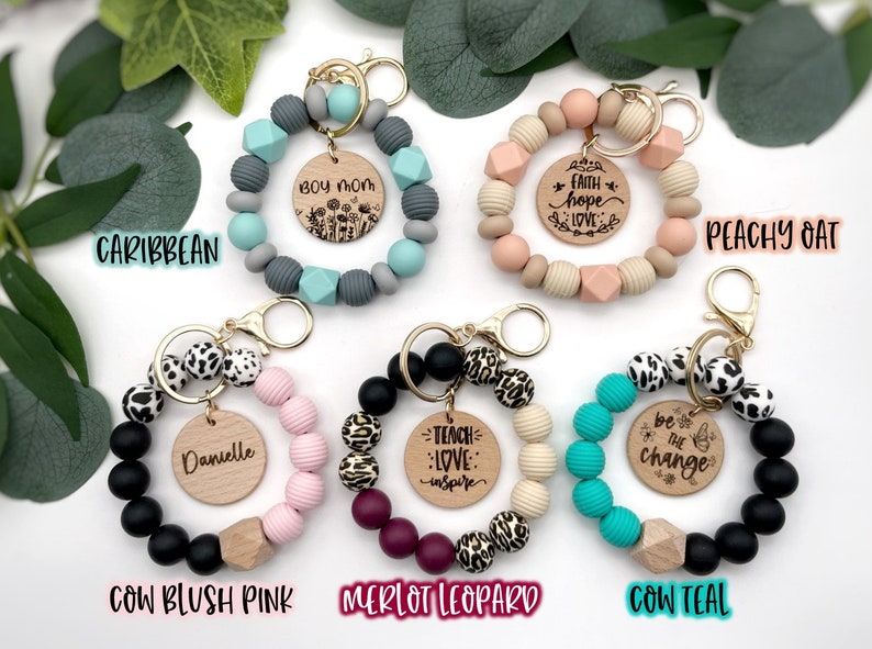 May include: Assortment of colorful beaded keychains with wooden charms. The charms feature various phrases, including "Boy Mom", "Faith Hope Love", "Teach Love Inspire", "Be the Change", and "Danielle".