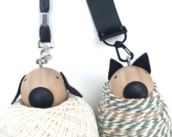 May include: Two wooden dog-shaped keyrings. One dog has black floppy ears and is wrapped in white yarn. The other dog has black pointed ears and is wrapped in green, brown, and white yarn. Both have black noses and are attached to black lanyards.