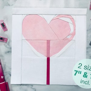 May include: A pink and white heart-shaped quilt block with a red stick, resembling a lollipop. The block is on a white background and is surrounded by white fabric. The text "2 sizes 7" & 12" incl." is written in green on a white circle.