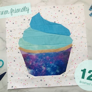 Könnte beinhalten: Ein Stoff-Cupcake-Quiltblock mit blauem und türkisfarbenem Zuckerguss-Design und einem Liner im Galaxien-Thema. Der Block ist 30,48 cm groß und mit "Anfängerfreundlich" beschriftet. Schere und andere Nähwerkzeuge sind sichtbar.