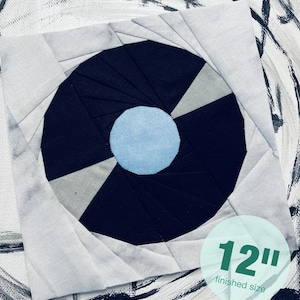 May include: A 12-inch quilt block with a black and white spiral design and a light blue circle in the center. The block is made of fabric and is finished with a white background.