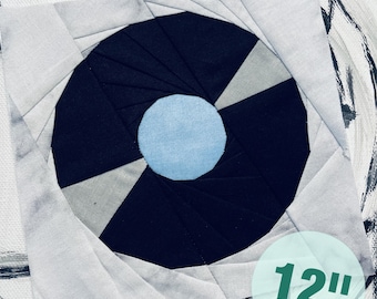 Könnte beinhalten: Ein 30,5 cm großer Quiltblock mit einem schwarzen und weißen Spiralmuster und einem hellblauen Kreis in der Mitte. Der Block ist aus Stoff gefertigt und hat einen weißen Hintergrund.