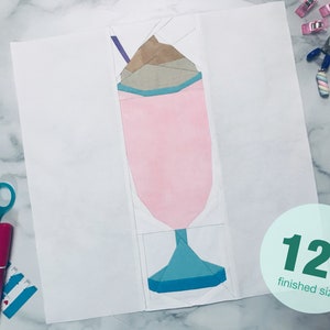 Puede incluir: Un patrón de papel para un bloque de colcha de batido de 30,5 cm de tamaño terminado. El patrón está formado por diferentes piezas de papel de colores que se recortan y pegan para crear el diseño. El batido es rosa con una cobertura de crema batida marrón y blanca y un vaso azul y verde.