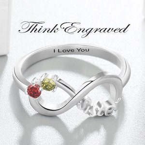 May include: Silver infinity ring with red and green gemstones. The ring is engraved with the word "Sister" and the phrase "I Love You".