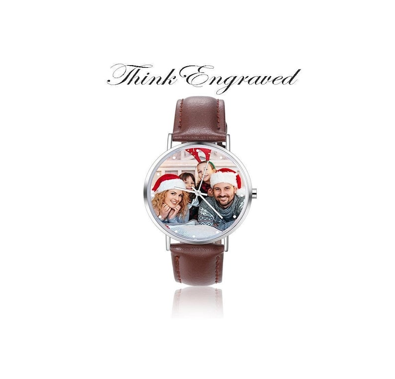 May include: A brown leather wristwatch with a silver face. The face of the watch has a photo of a family wearing Santa hats. The watch has the text "Think Engraved" above the watch.