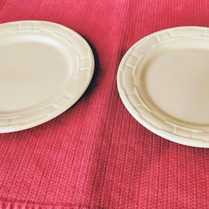 May include: Two round, cream-colored ceramic plates featuring a subtle, embossed geometric border pattern along the rim. The plates have a smooth, matte finish and a simple, clean design, presented side-by-side on a textured surface.