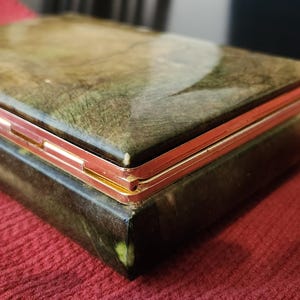 May include: A rectangular jewelry box with a marbled green and brown design on the lid and sides. The box has gold-colored metal trim and hinges, and it sits on a red fabric surface. The box is closed.