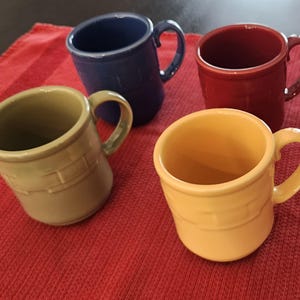 May include: Four ceramic mugs in blue, red, tan, and yellow. Each mug features a rounded handle and a textured, basket-weave pattern embossed around the middle section of the exterior. The mugs have a smooth, glossy finish and a consistent cylindrical shape.
