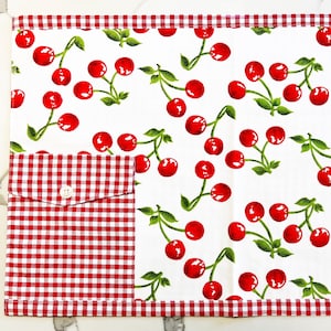 May include: Red and white checkered fabric placemat with a red gingham pocket and a single button. The placemat features a white background with a repeating pattern of red cherries with green stems and leaves.