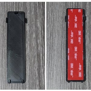 Puede incluir: Un objeto rectangular negro con una superficie texturizada. La parte posterior del objeto tiene una tira adhesiva roja con el texto "3M VHB" repetido varias veces.