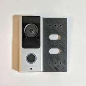 Op de afbeelding: Een zilveren en zwarte deurbelcamera met een zwarte lens en een knop. De deurbel is gemonteerd op een zwarte kunststof montageplaat met gaten voor schroeven.