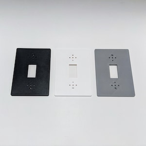 May include: Three light switch covers, one black, one white, and one grey. Each cover has a rectangular cutout for a light switch and a pattern of small holes around the edges.