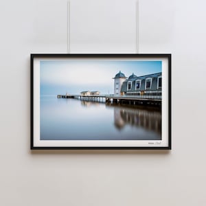 May include: A black and white photograph of a pier extending out into a calm body of water. The pier has a white building with a blue roof at the end. The sky is a light blue with some clouds. The water is a light blue and reflects the sky and the pier. The photo is framed in a black frame.