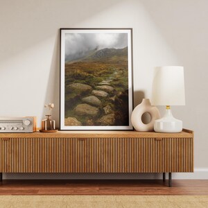 May include: A wooden media console with a light wood finish and a slatted front. The console has a stereo receiver, a lamp with a white shade, and a ceramic vase with dried flowers on top. A framed photograph of a mountain path with a stone walkway is hanging on the wall behind the console.