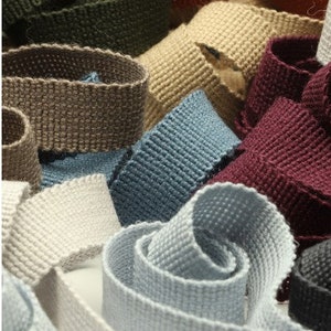 Può includere: Una collezione di nastri in tessuto tessuto in vari colori, tra cui beige, blu, grigio e bordeaux. Il nastro è testurizzato e ha una superficie leggermente ruvida.