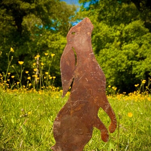 May include: A rusty metal silhouette of a rabbit standing tall in a field of green grass and yellow wildflowers. The rabbit is facing forward with its head tilted up.
