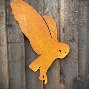 以下が含まれることがあります： 木製の壁に取り付けられた、さびたオレンジ色の金属製のフクロウのシルエット。フクロウは羽を広げて飛んでいます。