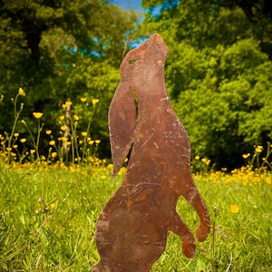 May include: A rusty metal silhouette of a rabbit standing tall in a field of green grass and yellow wildflowers. The rabbit is facing forward with its head tilted up.