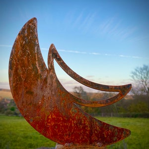 May include: A rusty metal crescent moon sculpture with a unique design. The sculpture is outdoors against a blue sky with clouds.