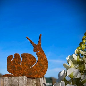 May include: A rusty metal snail silhouette decoratively mounted on a wooden fence post. The snail is set against a bright blue sky, with white flowers in the background. A whimsical garden accent.