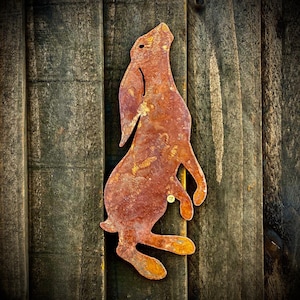 May include: A rusty metal cutout of a rabbit or hare, standing on its hind legs with its ears up, mounted on a wooden surface.