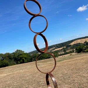 May include: A rusty metal sculpture of five interlocking circles, arranged vertically, against a backdrop of a blue sky and green hills.