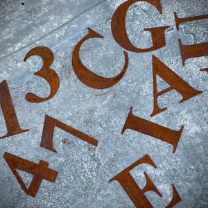 May include: Rusty metal letters and numbers scattered on a textured gray surface. The letters include G, A, P, and E. Numbers include 1, 3, 4, and 8. The metal has a weathered, aged appearance.