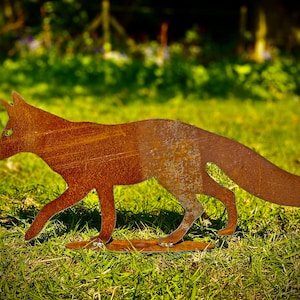 Puede incluir: Una decoración de jardín de metal oxidado en forma de silueta de zorro. El zorro camina con la cola levantada. El zorro está de pie sobre un césped verde.