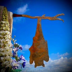 Op de afbeelding: Een roestig metalen silhouet van een vleermuis hangend aan een tak tegen een blauwe lucht. De vleermuis hangt ondersteboven met zijn vleugels uitgespreid.
