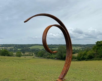 Puede incluir: Una escultura de metal oxidado en forma de un gran remolino abstracto. La escultura está de pie en un campo con una colina verde ondulada en el fondo.