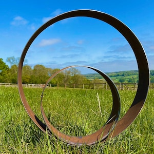 May include: Two large rusty metal rings, one inside the other, stand in a grassy field. The rings are positioned to frame a view of a rolling green hillside and a blue sky with white clouds.