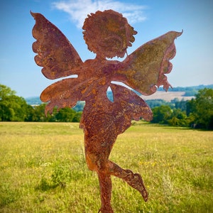 May include: A rusty metal fairy sculpture with outstretched wings, standing in a field of green grass. The fairy is facing to the right and has a whimsical, stylized design.
