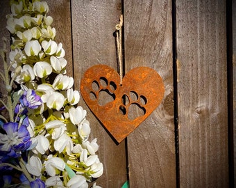 May include: A rusty metal heart-shaped ornament with two paw prints cut out. The ornament is hanging from a string on a wooden background.