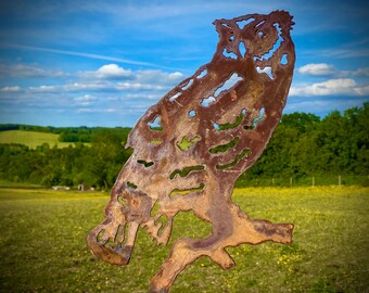 May include: A rusty metal owl sculpture with a cutout design. The owl is perched on a branch and has a detailed face with large eyes. The sculpture is set against a backdrop of a green field and a blue sky with white clouds.