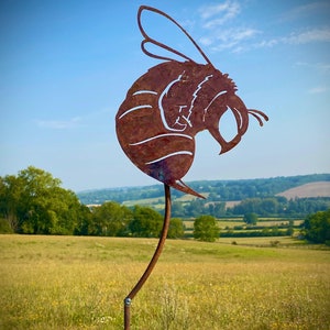 May include: A rusty brown metal bee sculpture with a long, thin stem, standing in a field of long grass. The bee is facing to the right and has a detailed design.