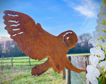 May include: A rusty metal owl silhouette with outstretched wings mounted on a wooden fence post. The owl is facing right and is in front of a green field and a blue sky with white clouds.