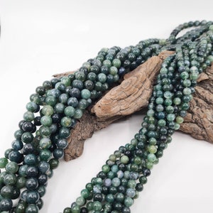 May include: A strand of green agate beads, with a mottled green and blue colour, arranged on a piece of driftwood. The beads are round and smooth, and they have a natural, earthy look.